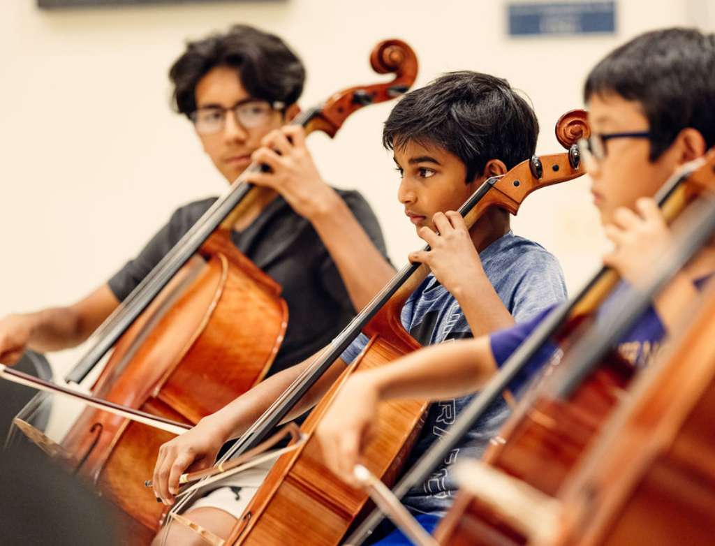 A group of three cello students play their instruments together and look at each other to coordinate their timing.
