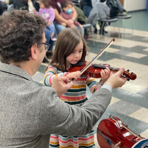 An adult kneels down to help a young child hold a violin and bow.