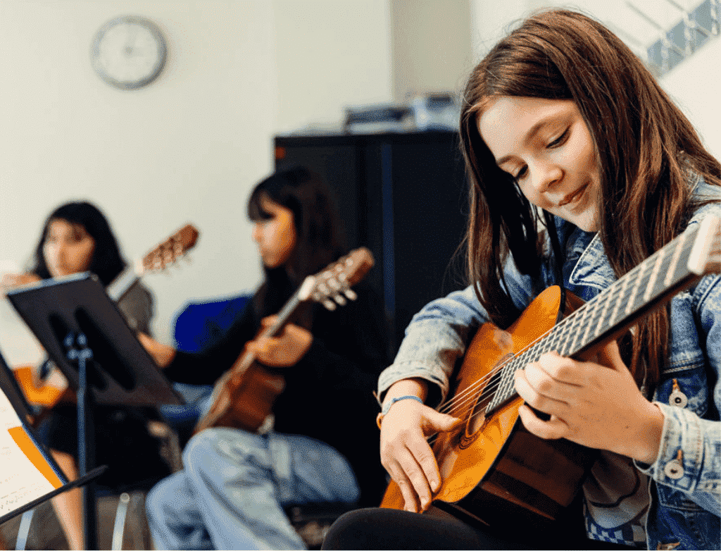 A young student in the foreground smiles down at the guitar she's playing. Two other students are blurred in the background.