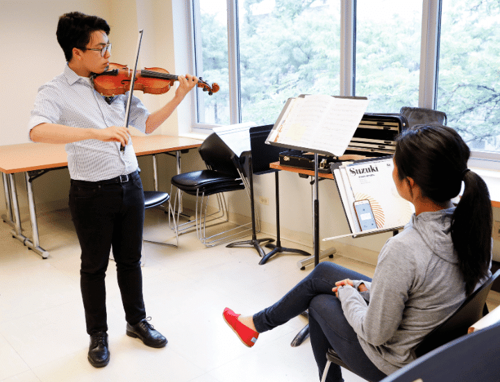 A student stands playing the violin in front of his teacher.